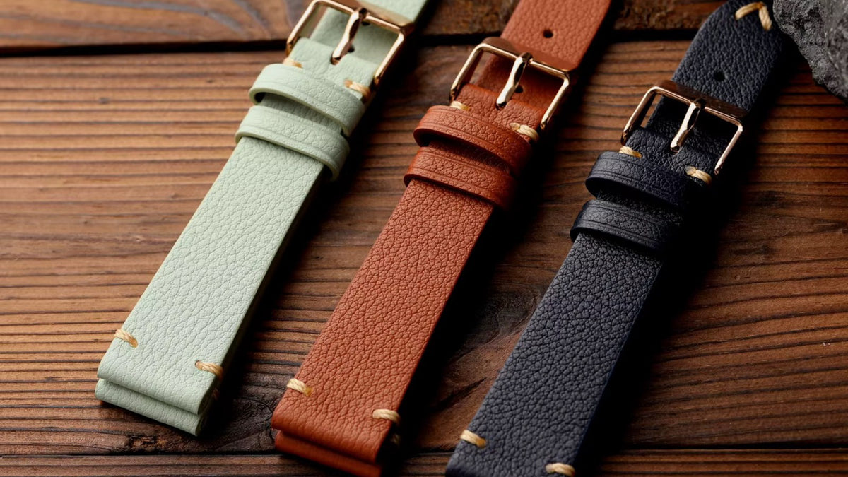 Sage green, tan and navy goat leather watch straps on a dark wood surface